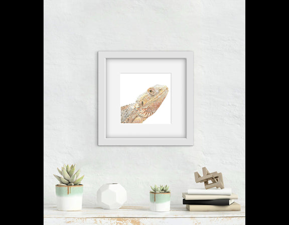 Framed artwork of a bearded dragon lizard on a white wall with decorative items below.