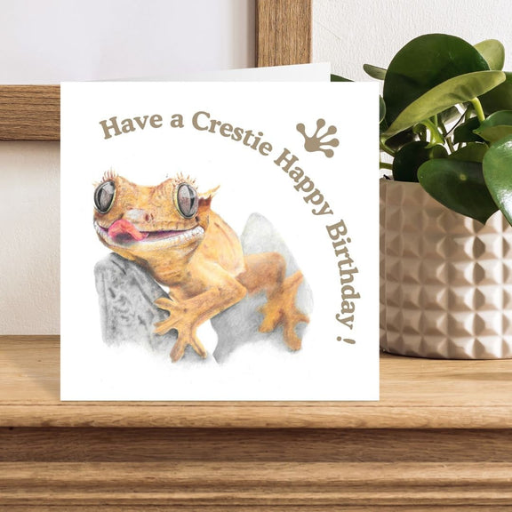 Birthday card with a crested gecko illustration on a wooden surface with a plant in the background.