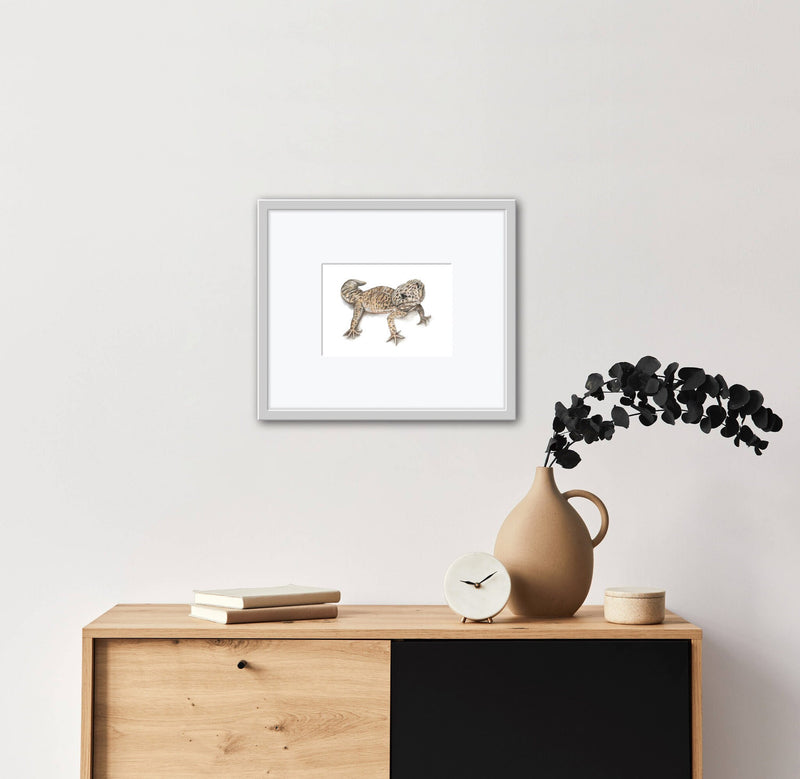 Framed artwork of a Leopard Gecko on a wall above a wooden cabinet with decor items.