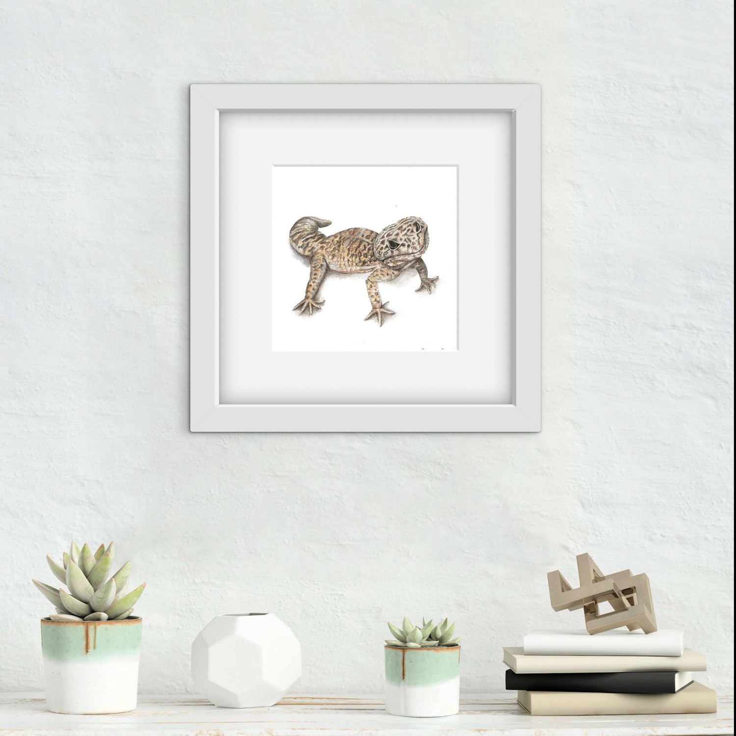 Framed artwork of a drawing of a Leopard Gecko Lizard on a white wall with decorative items below.