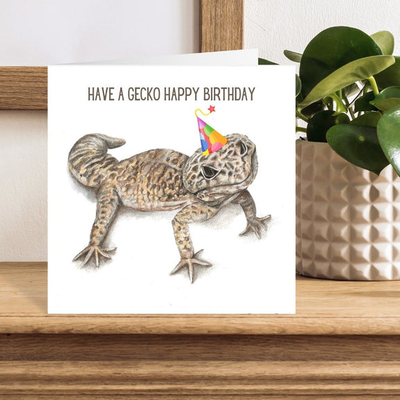Birthday card with a gecko wearing a party hat on a wooden surface with a plant in the background.
