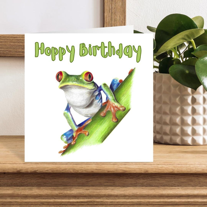 Birthday card with a frog illustration on a wooden surface with a plant in the background