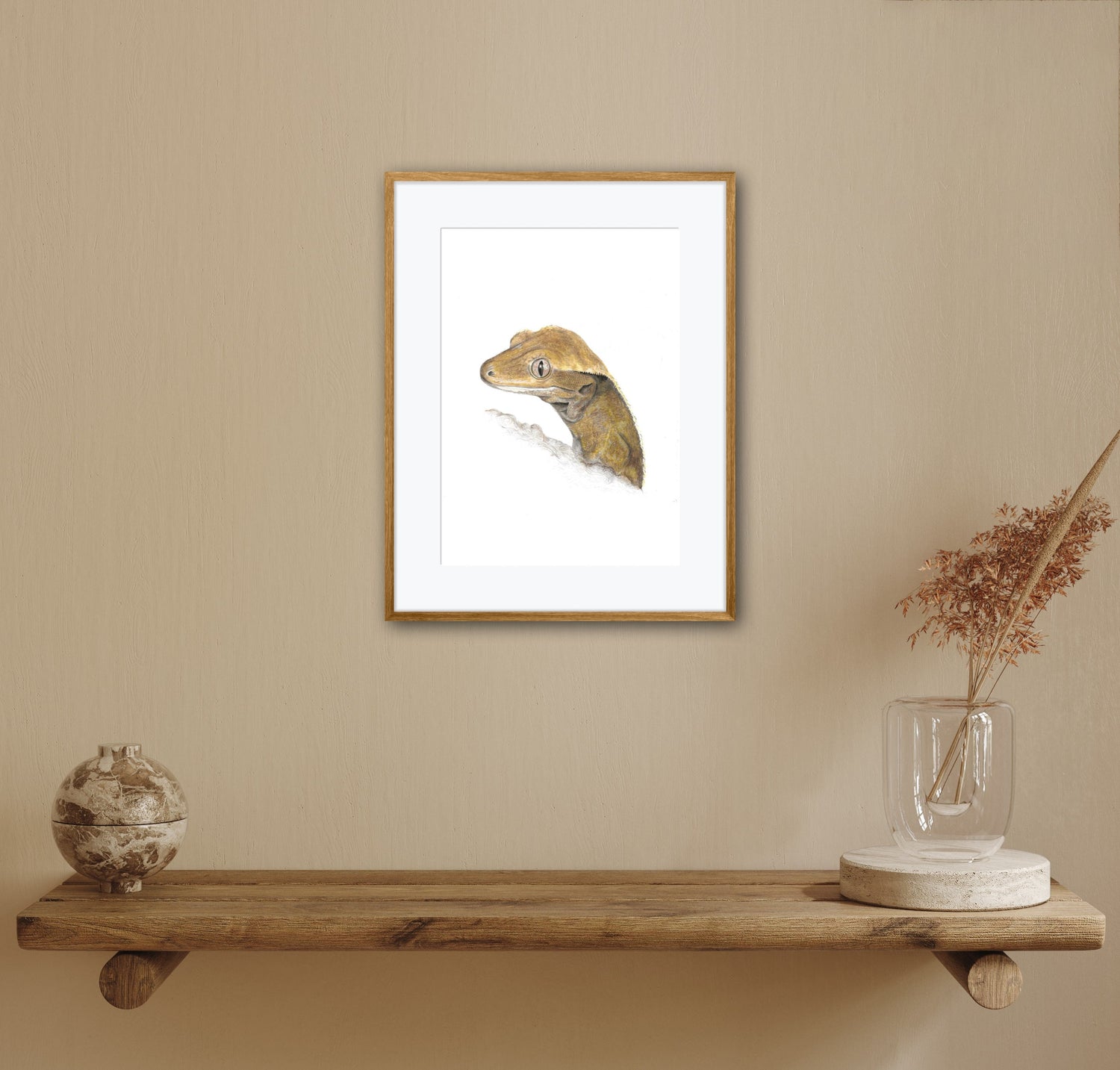 Framed artwork of a crested gecko reptile on a beige wall above a wooden shelf with decorative items.