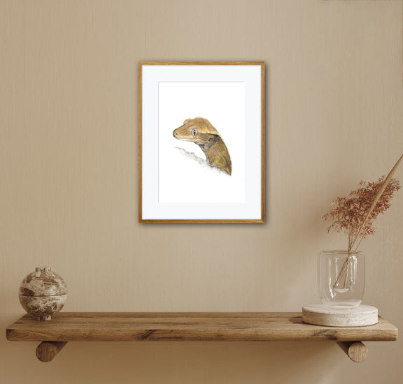 Framed artwork of a crested gecko reptile on a beige wall above a wooden shelf with decorative items.