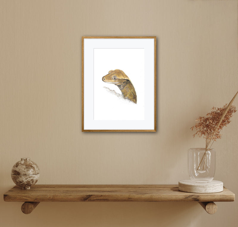 Framed artwork of a crested gecko reptile on a beige wall above a wooden shelf with decorative items.