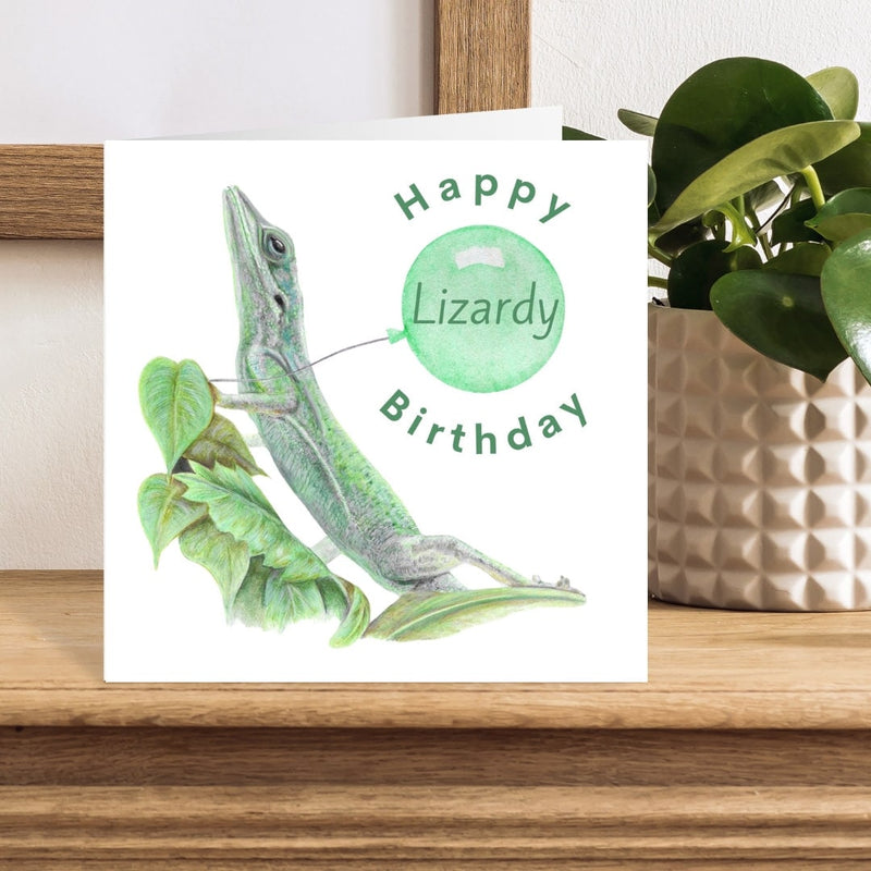 Birthday card with a green lizard illustration and 'Happy Lizardy Birthday' text on a wooden surface.
