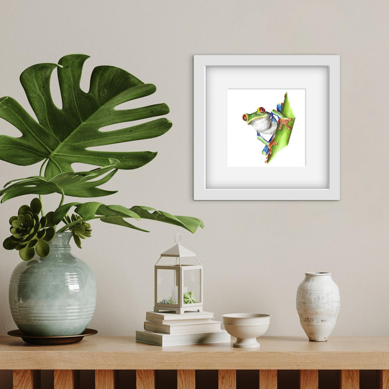 Framed colourful frog artwork on a wall with decorative items on a table below.