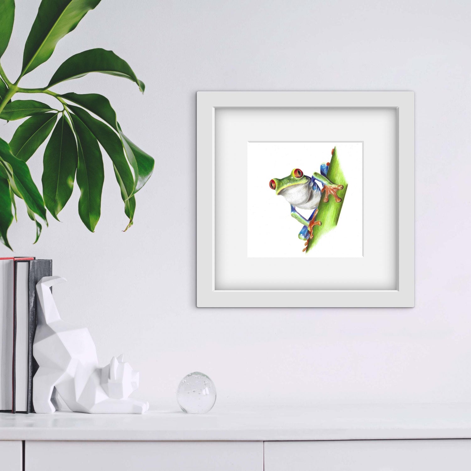 Framed artwork of a frog on a leaf with a plant and books on the shelf below
